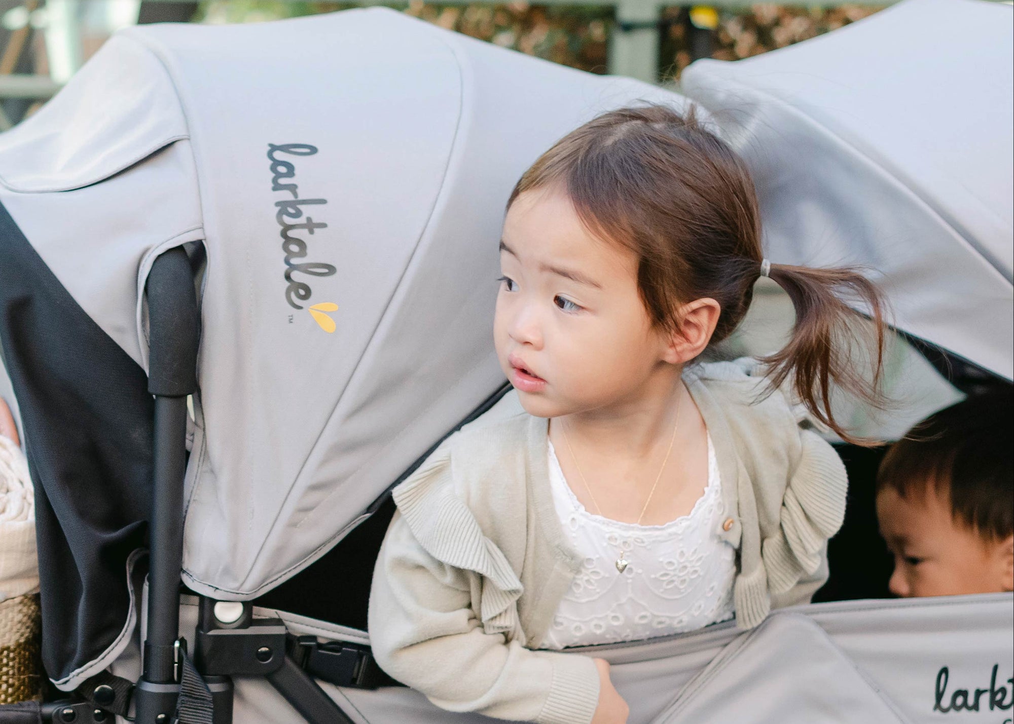 Toddler girl peeks out from under the sun canopy of the caravan stroller wagon in gray and black